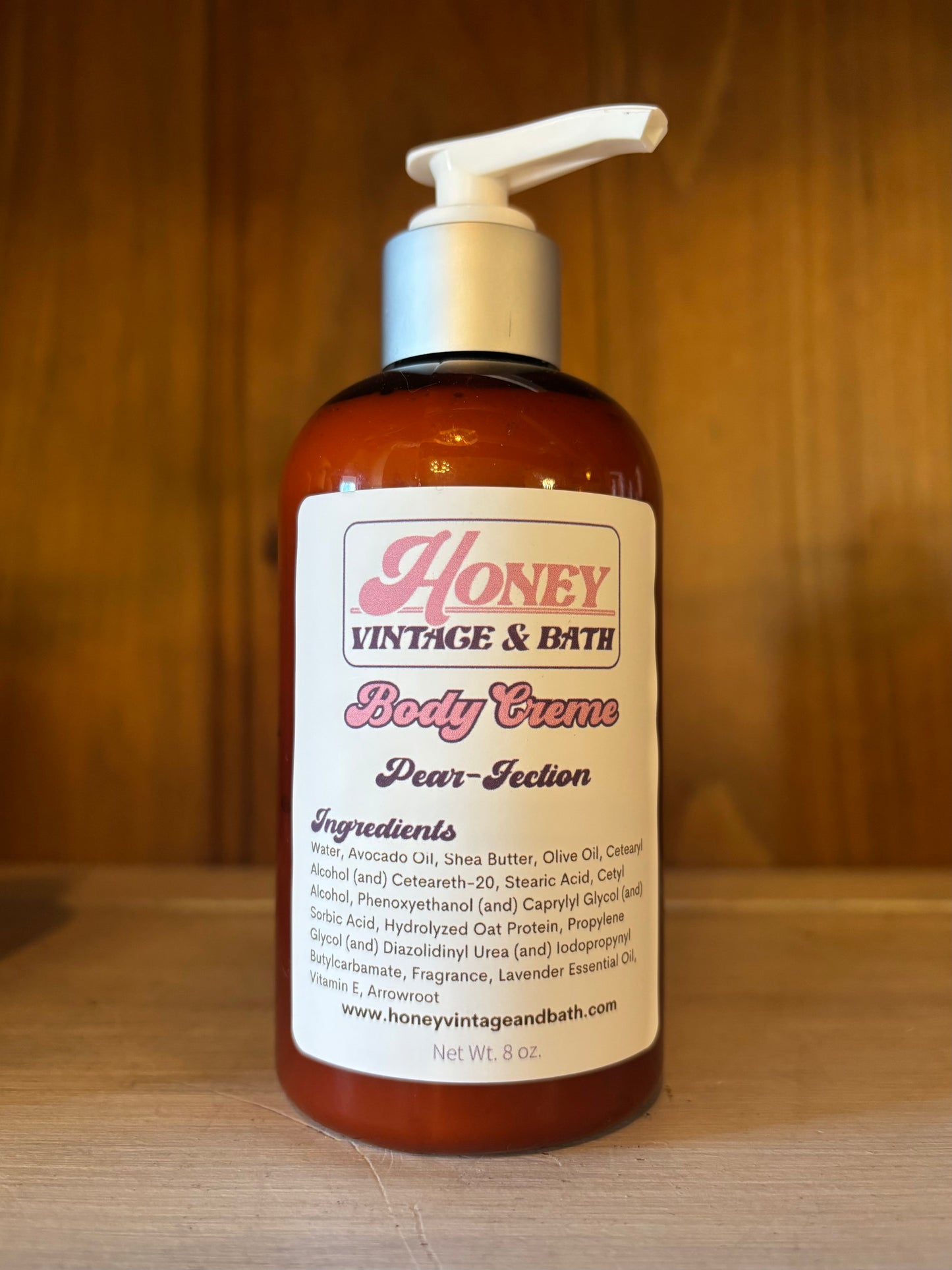 A brown plastic bottle with a white pump dispenser contains “Honey Vintage & Bath Body Creme” in the “Pear-fection” scent. The label lists ingredients like water, avocado oil, shea butter, olive oil, with a net weight of 8 fl. oz. The background is a wooden surface.