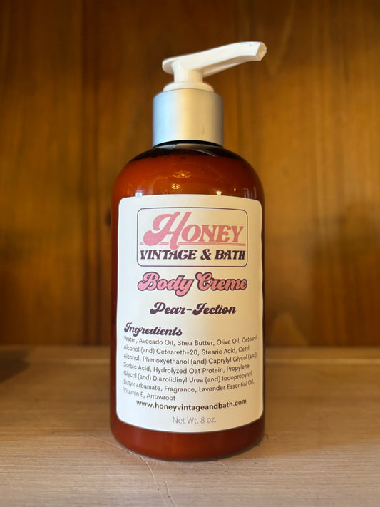 A brown plastic bottle with a white pump dispenser contains “Honey Vintage & Bath Body Creme” in the “Pear-fection” scent. The label lists ingredients like water, avocado oil, shea butter, olive oil, with a net weight of 8 fl. oz. The background is a wooden surface. 