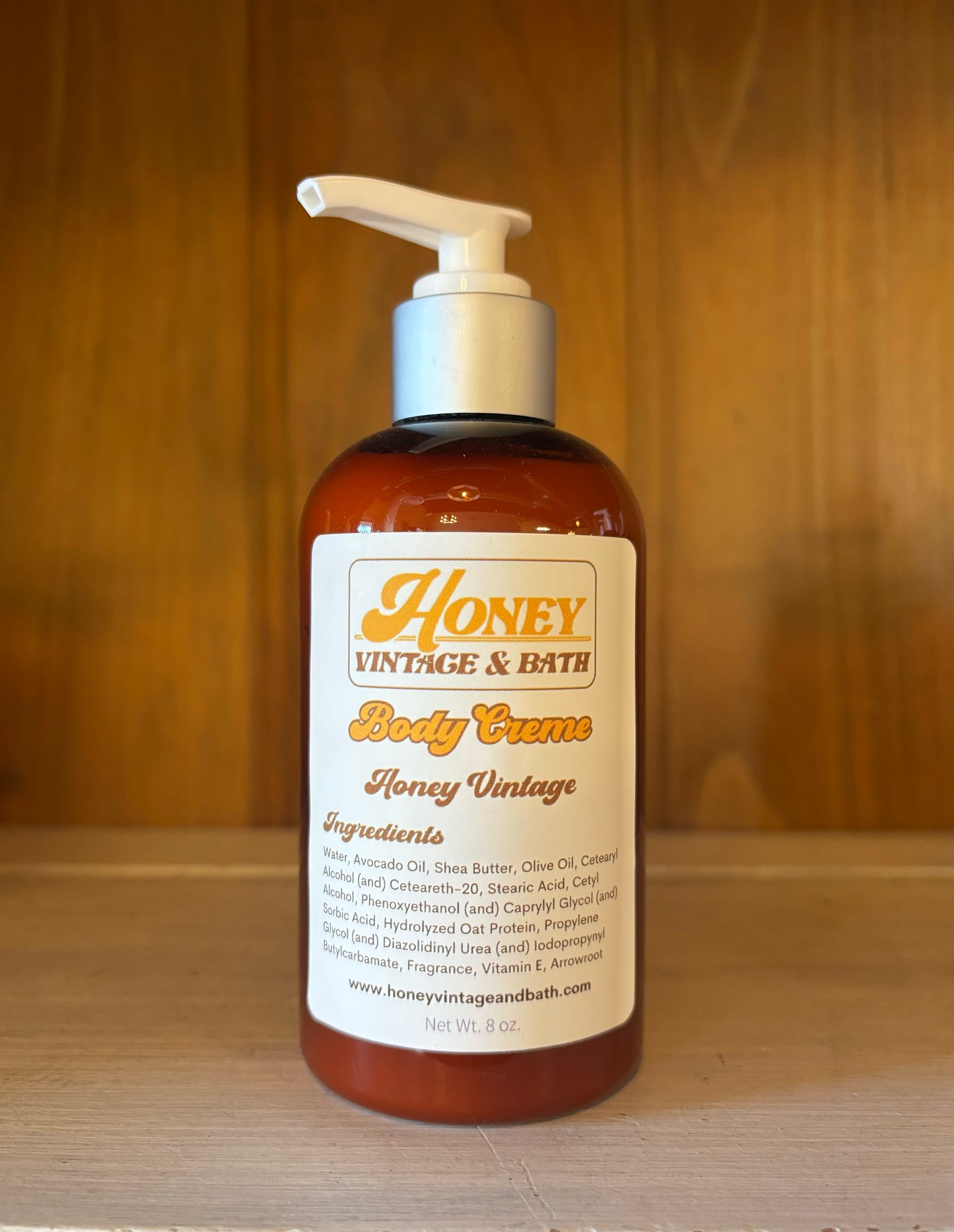 A brown plastic bottle with a white pump dispenser contains “Honey Vintage & Bath Body Creme” in the “Mango Tango” scent. The label lists ingredients like water, avocado oil, shea butter, olive oil, with a net weight of 8 fl. oz. The background is a wooden surface. 