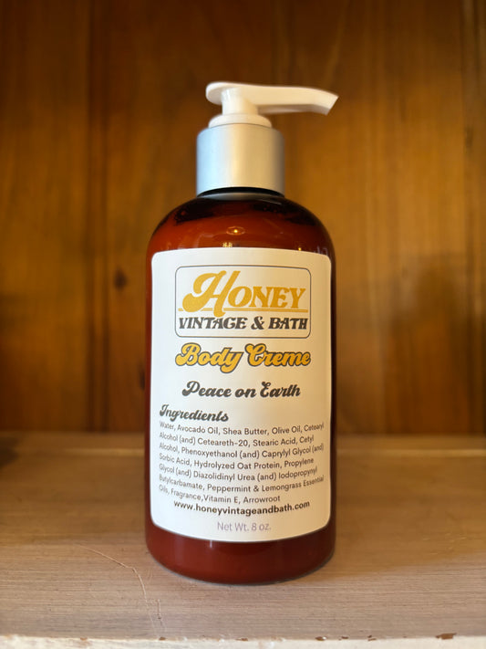 A brown plastic bottle with a white pump dispenser contains “Honey Vintage & Bath Body Creme” in the “Peace on Earth” scent. The label lists ingredients like water, avocado oil, shea butter, olive oil, with a net weight of 8 fl. oz. The background is a wooden surface. 