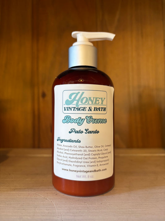 A brown plastic bottle with a white pump dispenser contains “Honey Vintage & Bath Body Creme” in the “Palo Santo” scent. The label lists ingredients like water, avocado oil, shea butter, olive oil, with a net weight of 8 fl. oz. The background is a wooden surface. 